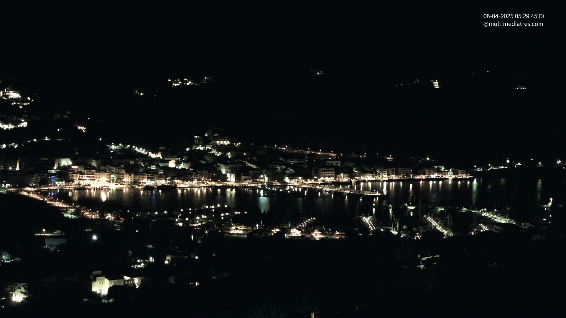 A nighttime view of a coastal town with many lights illuminating buildings and a harbor filled with boats, under a clear, dark sky.