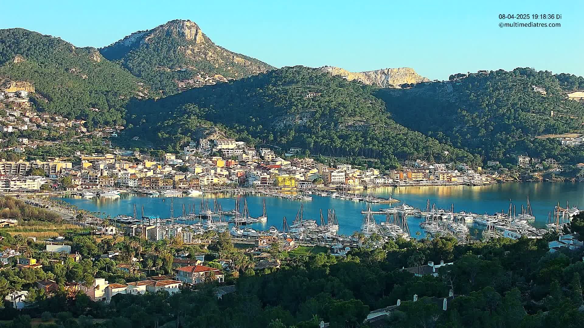 A coastal town nestled in a mountainous region, with a harbor full of sailboats under a clear, sunny sky.