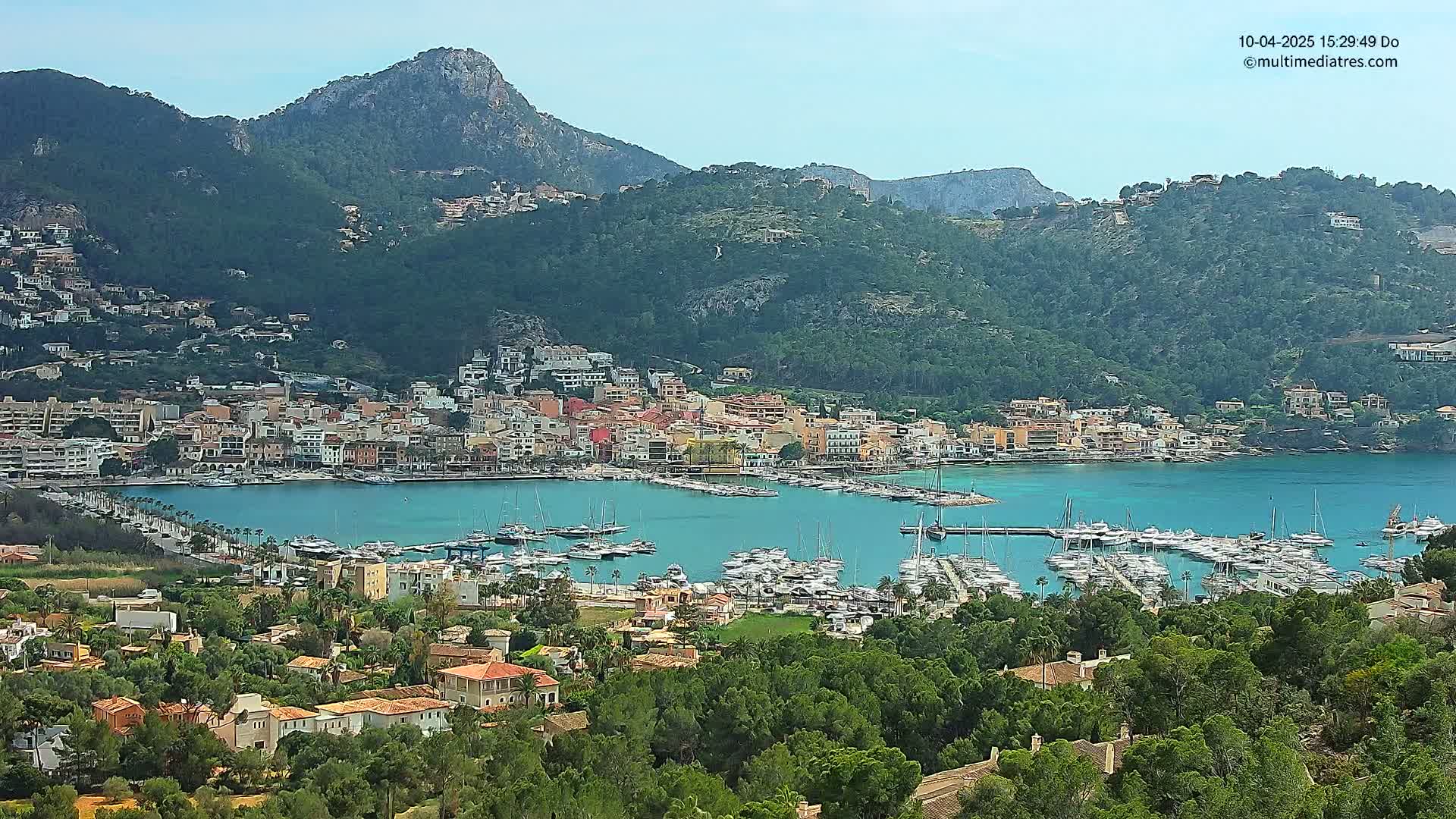 A picturesque coastal town nestled in a mountainous region, filled with docked boats in a calm, clear harbor under a sunny sky.