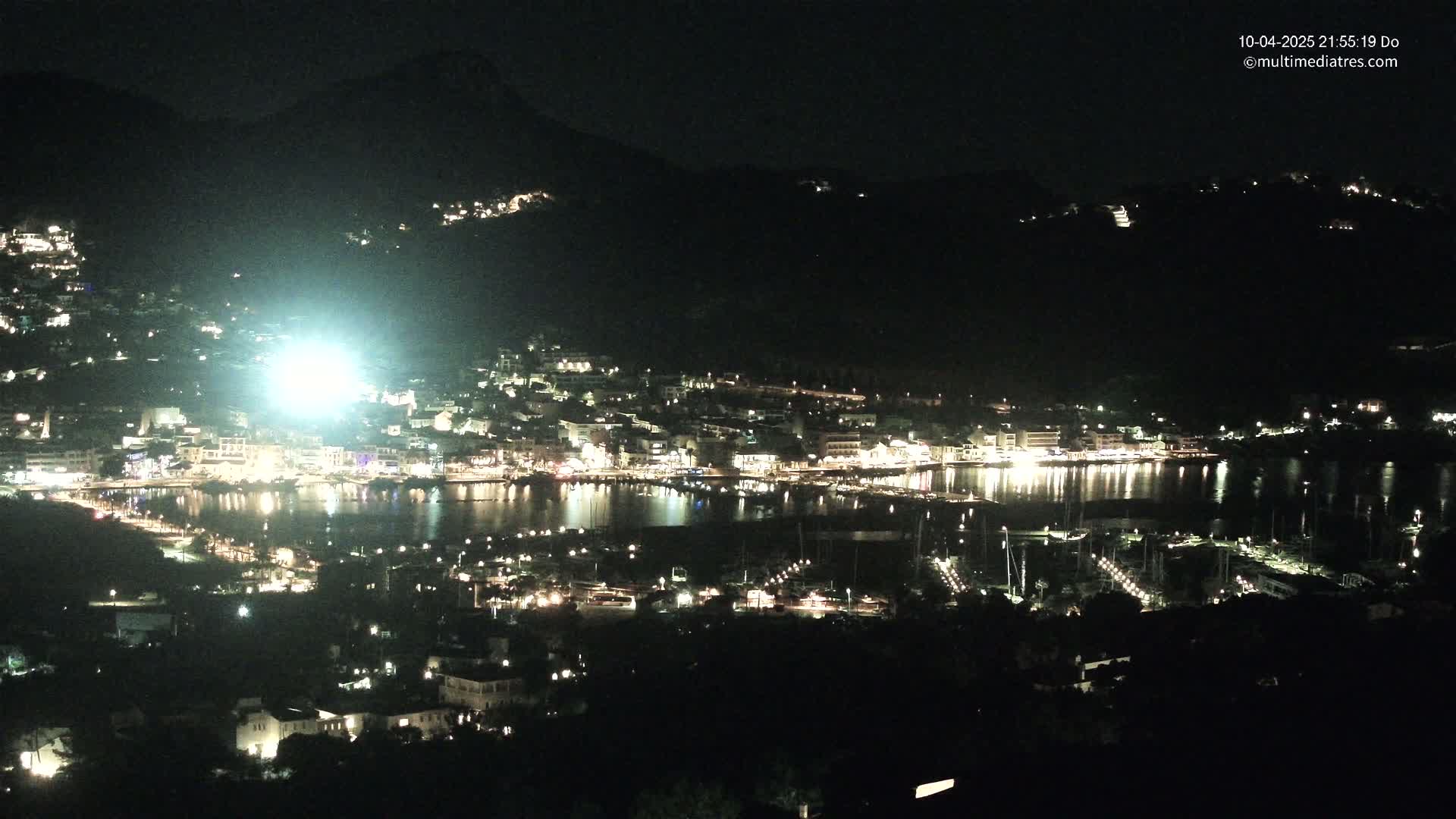 A nighttime, aerial view of a harbor town nestled between dark hills, with its lights reflected on the calm water.