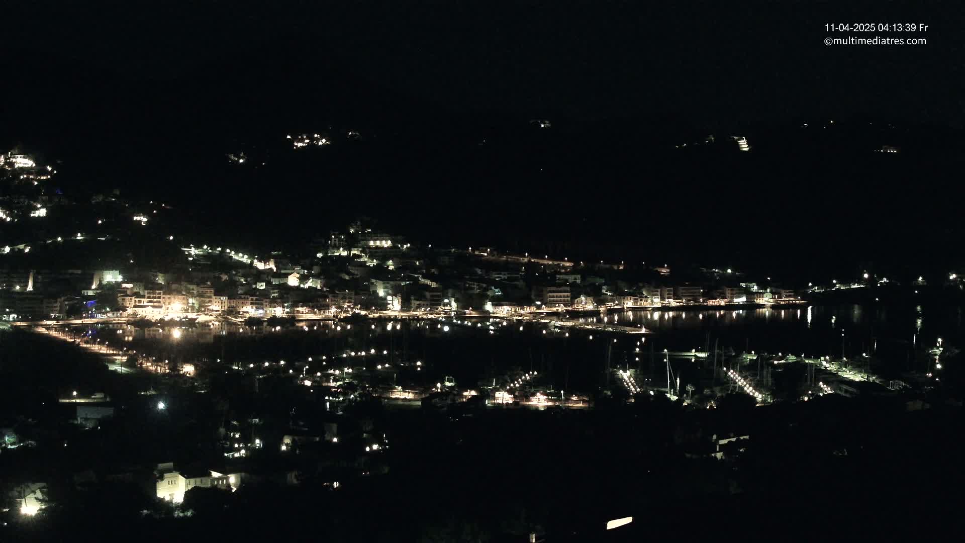 A coastal town at night, illuminated by lights from buildings and boats in a calm harbor.