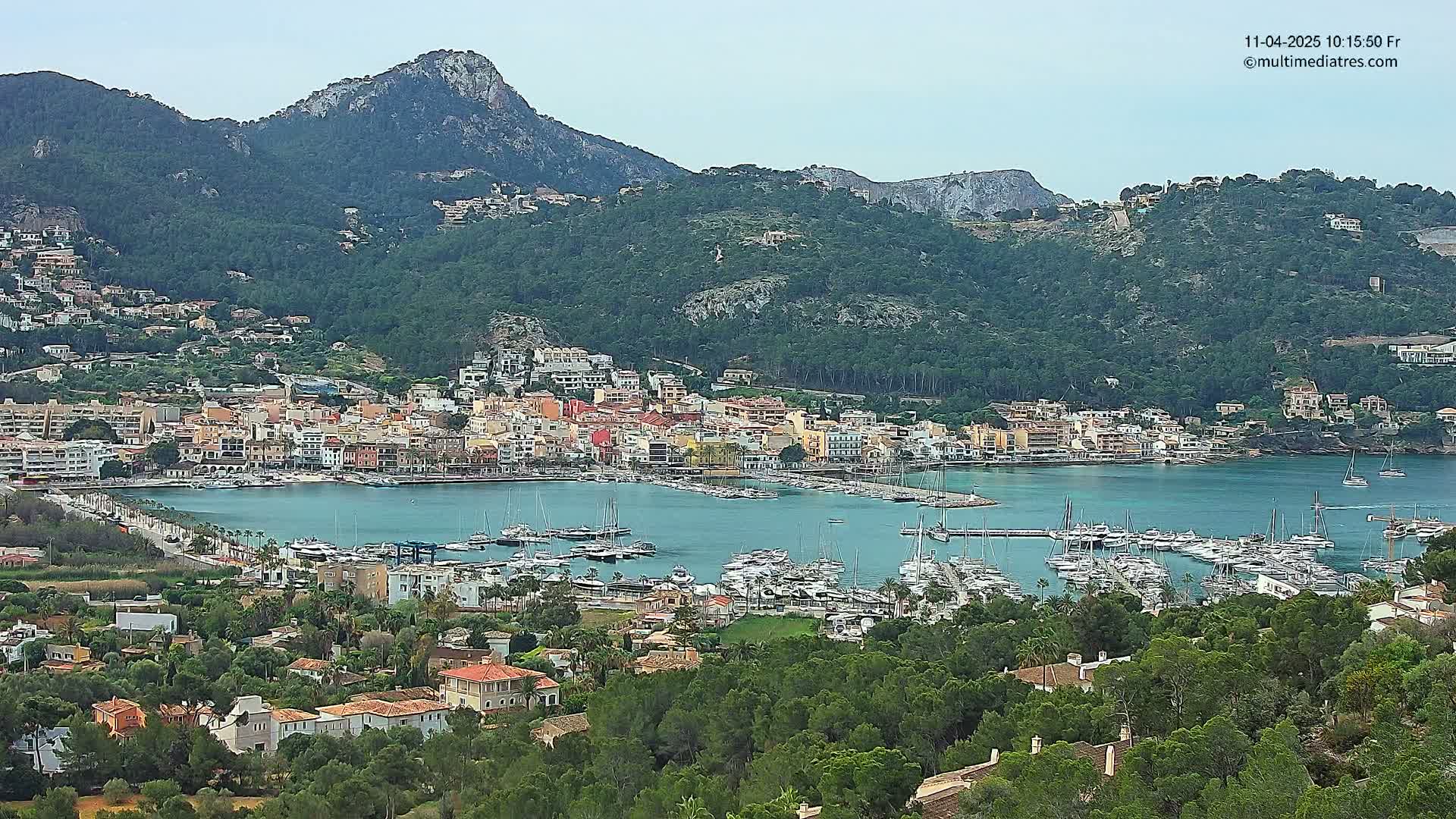 A coastal town with a marina full of sailboats is nestled between lush green hills under a mostly clear sky.