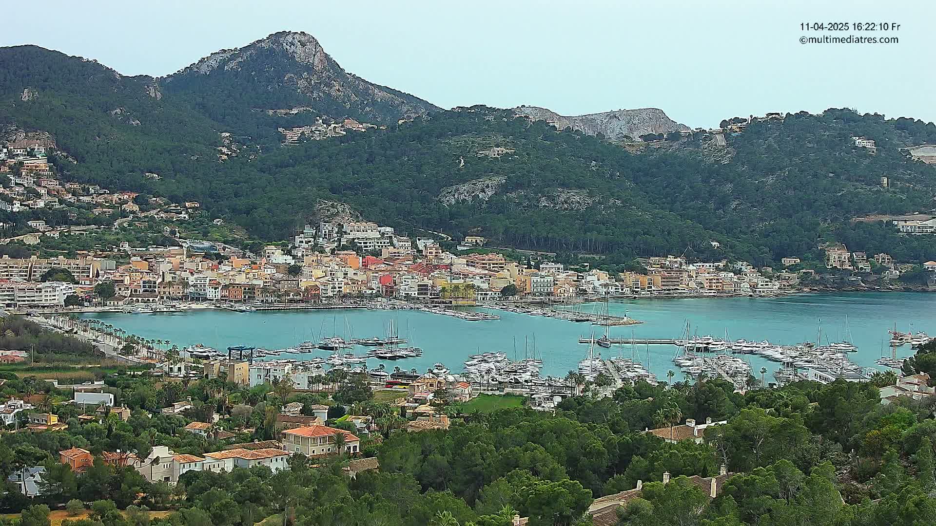 A coastal town with a marina filled with boats is nestled against a mountainside under a cloudy sky.