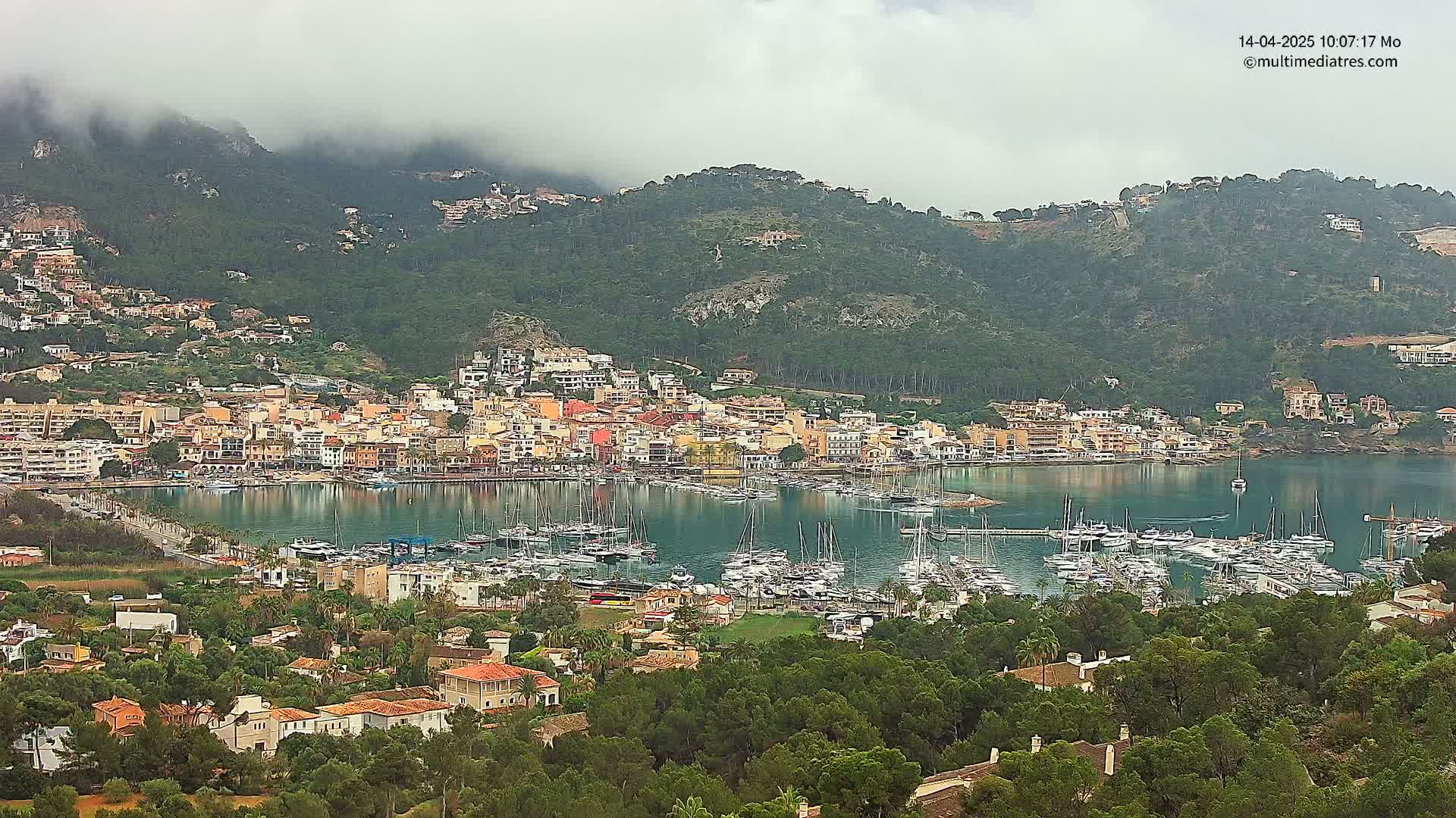 A coastal town nestled in a bay filled with sailboats, is situated beneath a mountain shrouded in fog.