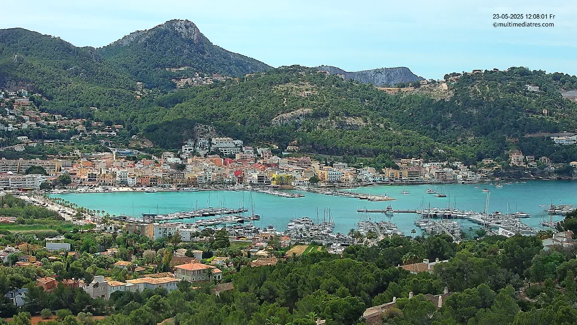 A picturesque coastal town nestled in a mountainous region, with a calm bay filled with numerous boats and yachts under a partly cloudy sky.