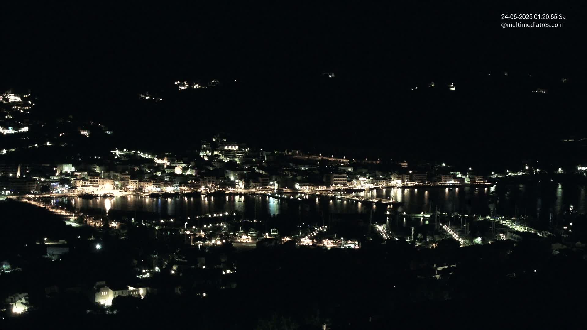 A nighttime view of a harbor town illuminated by lights, nestled along a dark shoreline under a clear, starless sky.