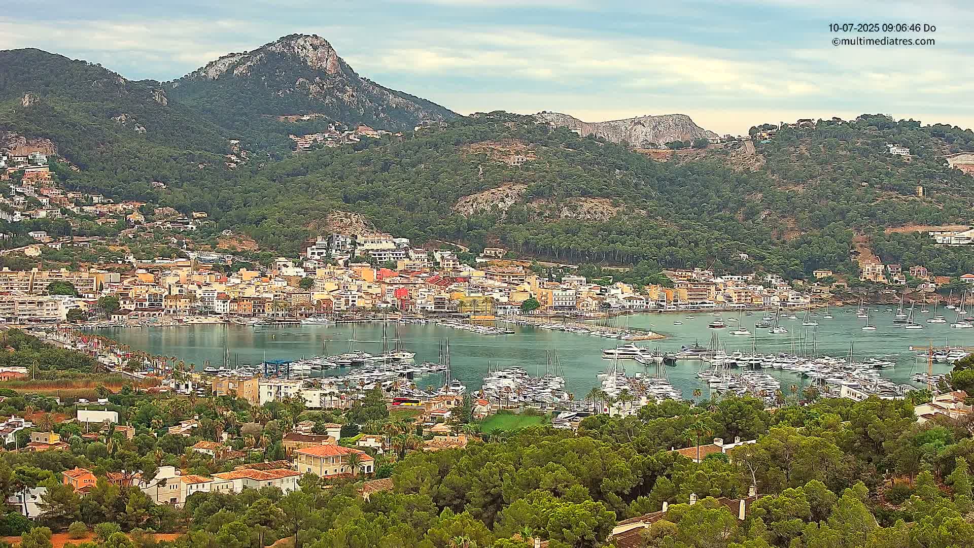 A coastal town nestled in a mountainous region, with a harbor full of boats under partly cloudy skies.