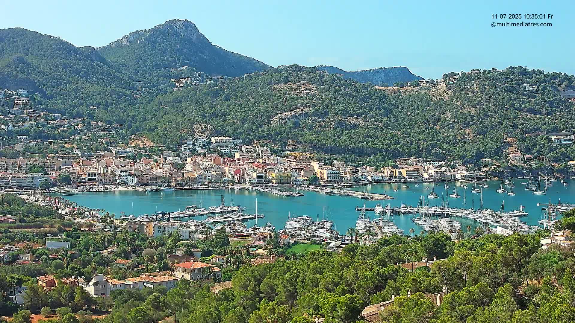 A coastal town nestled between green hills under a clear blue sky, with a harbor filled with numerous boats.
