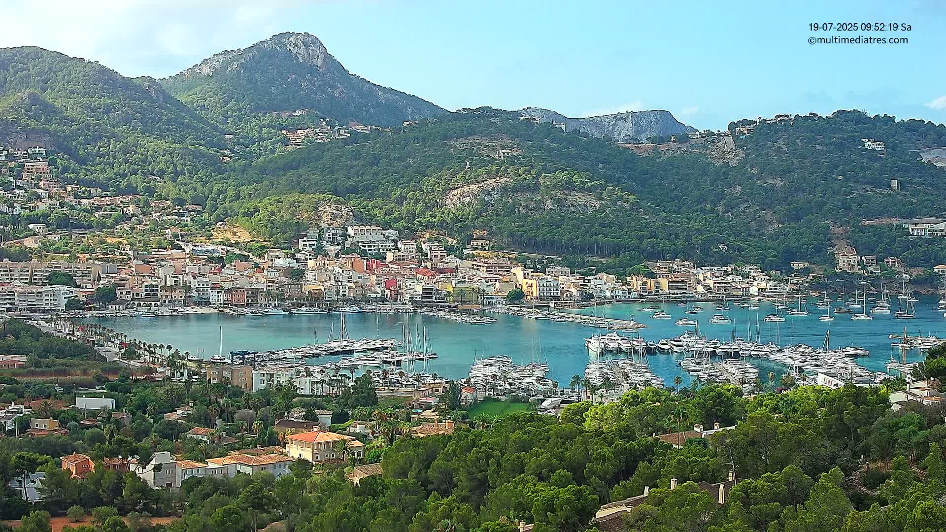 A picturesque coastal town nestled between lush green hills and a calm harbor filled with numerous boats on a sunny day.
