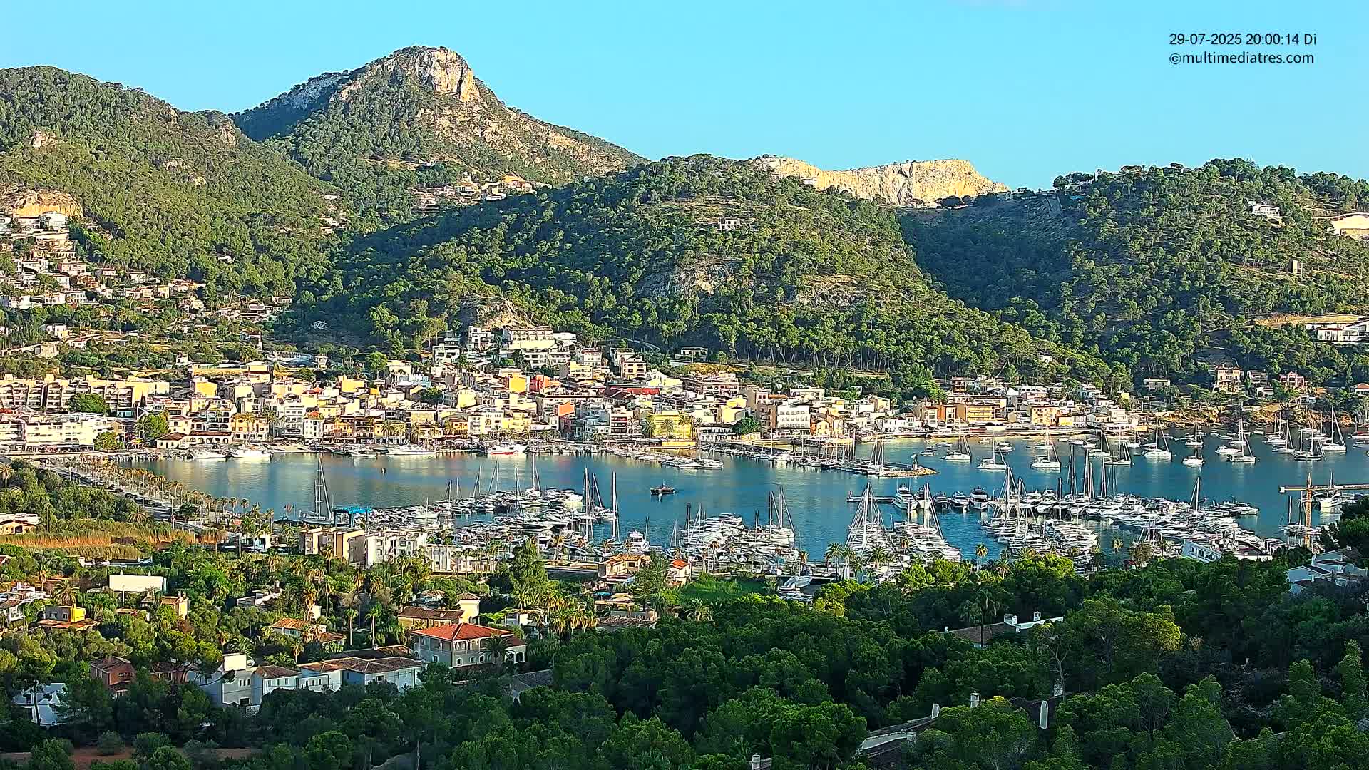 A coastal town nestled in a green mountain valley, filled with docked sailboats under a clear sky, overlooks a calm bay.