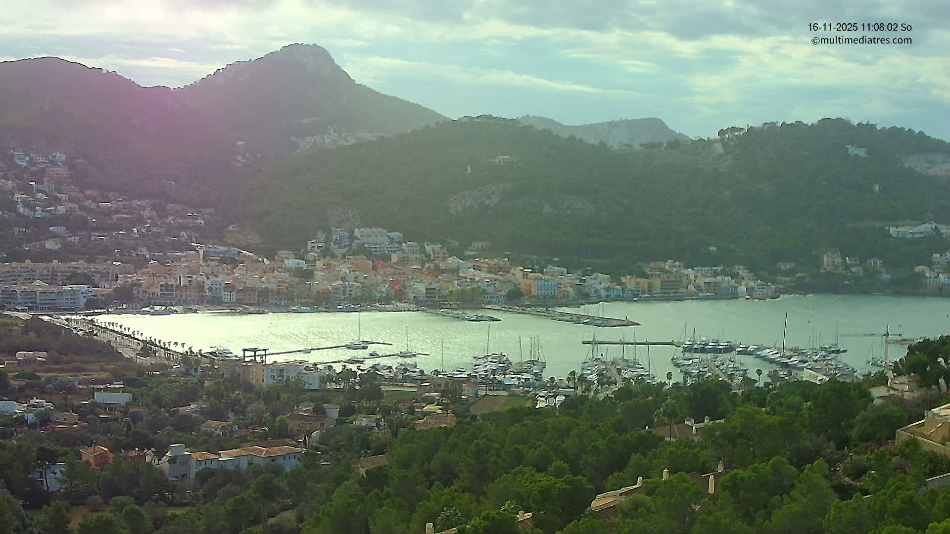 An elevated view reveals a picturesque coastal town with colorful buildings nestled around a tranquil bay filled with numerous moored boats, all set against a backdrop of verdant mountains under an overcast sky with diffused sunlight.