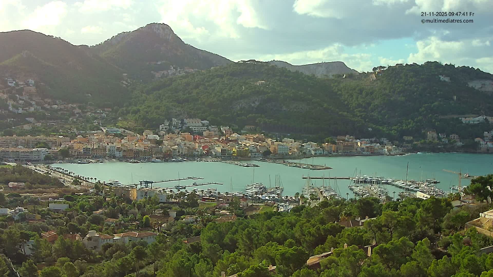 An elevated view reveals a picturesque coastal town with colorful buildings nestled around a busy harbor filled with boats, all set against a backdrop of verdant mountains under a partly cloudy sky.