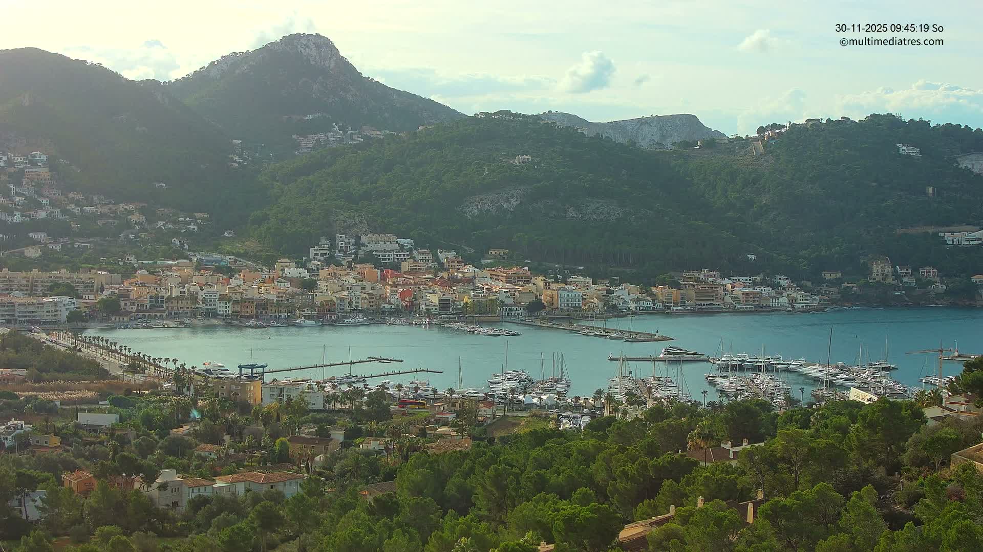 A vibrant coastal town with colorful buildings and numerous boats docked in its calm bay is nestled amidst lush green mountains under a partly cloudy and bright sky.