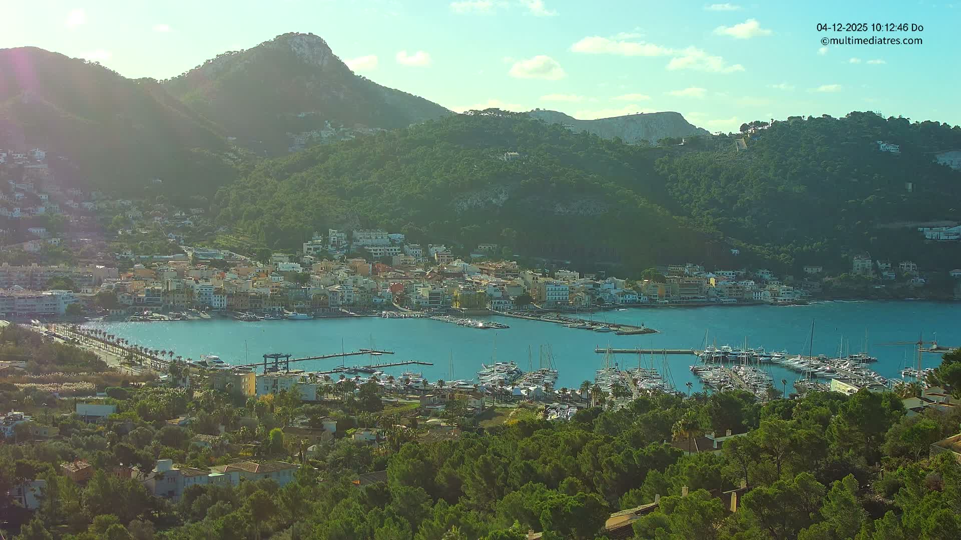 A vibrant coastal town featuring a busy marina filled with boats is nestled in a calm bay, surrounded by verdant mountains under a bright, partly cloudy sky.