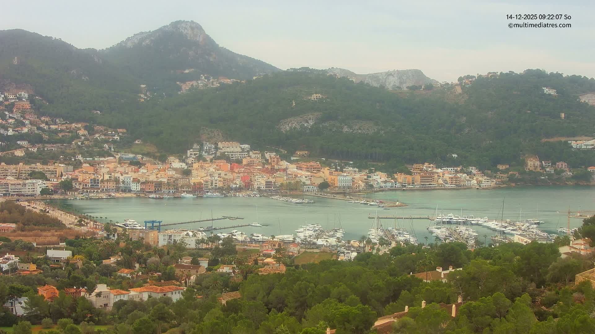 A vibrant coastal town featuring a busy marina filled with boats is nestled in a calm bay, surrounded by verdant mountains under a bright, partly cloudy sky.