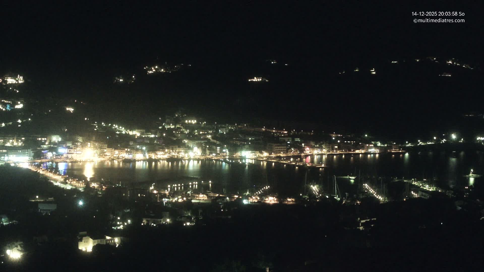 A vibrant coastal town featuring a busy marina filled with boats is nestled in a calm bay, surrounded by verdant mountains under a bright, partly cloudy sky.