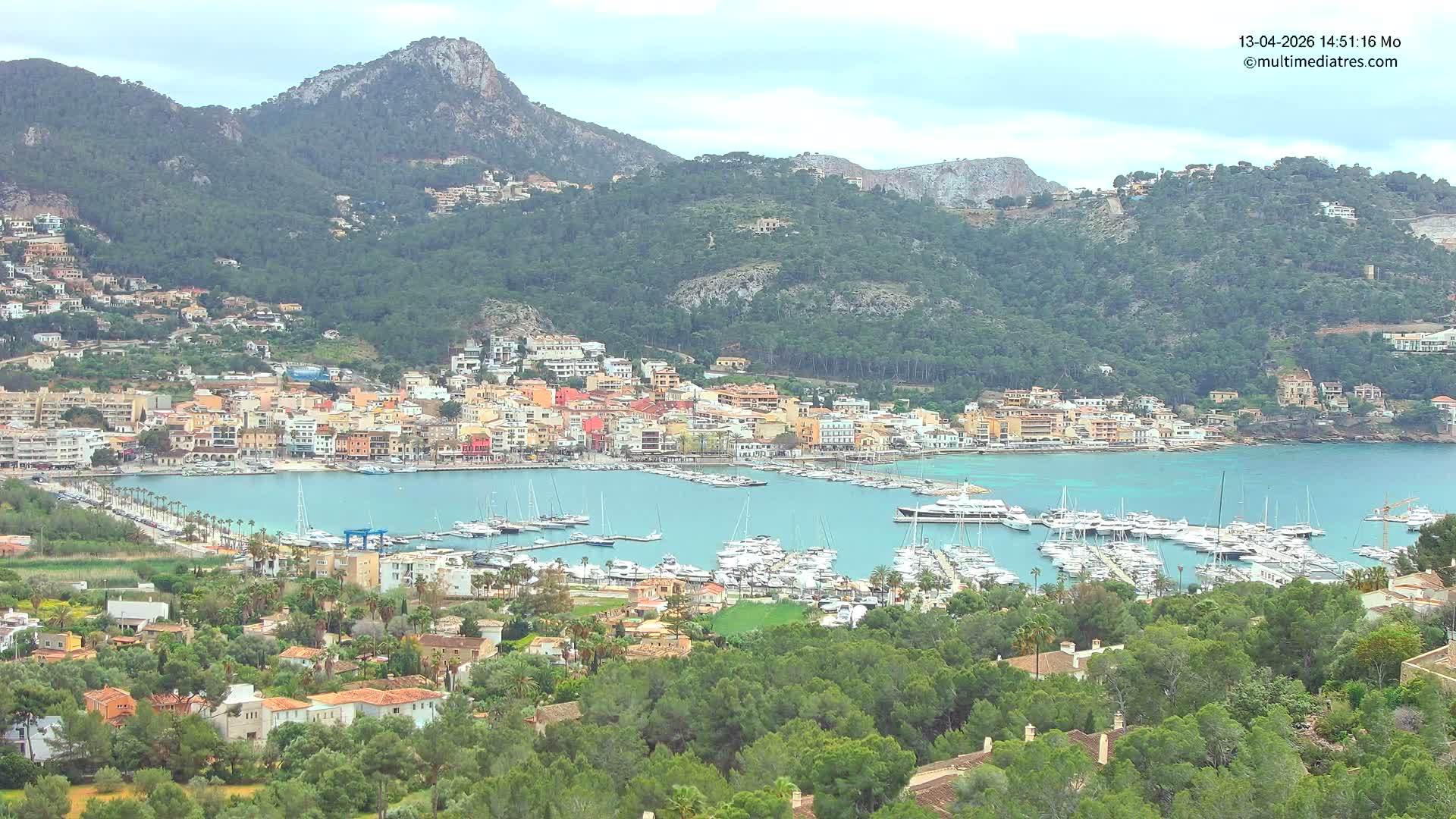 A vibrant coastal town featuring a busy marina filled with boats is nestled in a calm bay, surrounded by verdant mountains under a bright, partly cloudy sky.