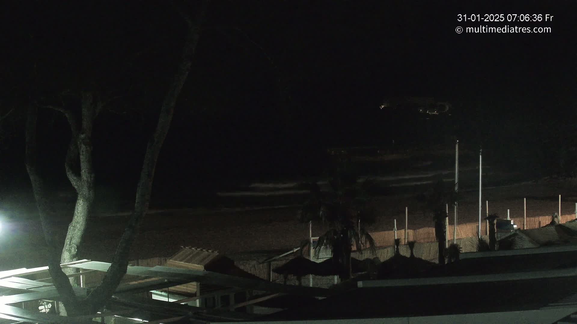 A dark nighttime scene shows a beach with calm waves, some palm trees, and beach huts under a mostly clear sky.