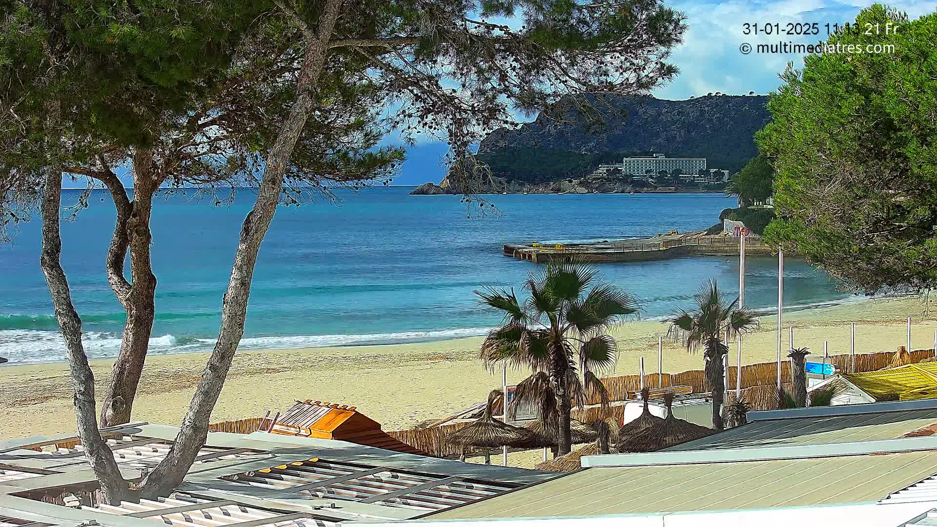 A sandy beach with calm, blue water, palm trees, and buildings is seen from an elevated viewpoint on a partly sunny day.