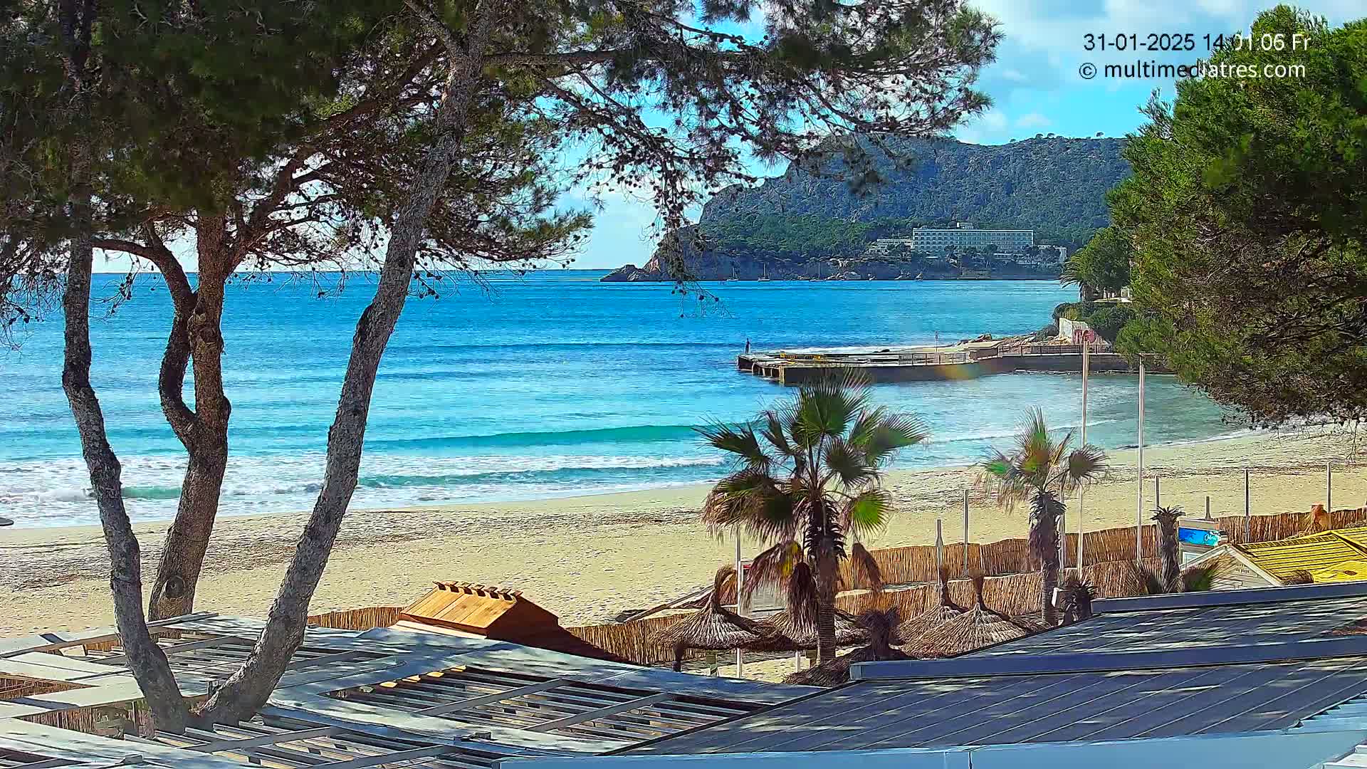 A sandy beach with turquoise water, gentle waves, palm trees, and beachside structures is visible under a mostly sunny sky.