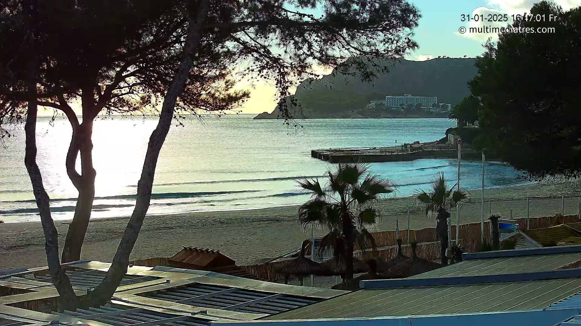 A sandy beach with gentle waves laps the shore under a partly cloudy sky, framed by trees and buildings, with a calm sea stretching to a distant hillside.