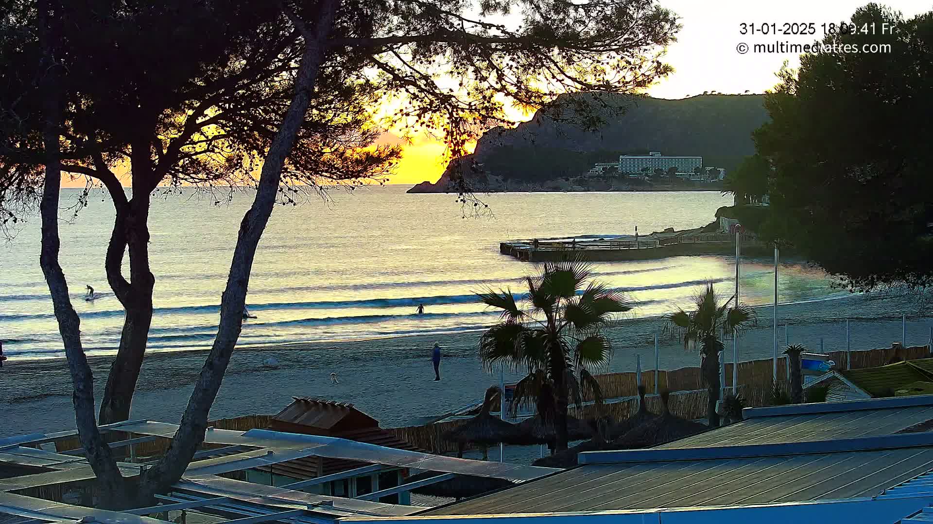 A sandy beach with gentle waves laps the shore at sunset, under a clear sky, with a few people visible on the beach and in the water, behind which sits a hotel and a rocky hill.