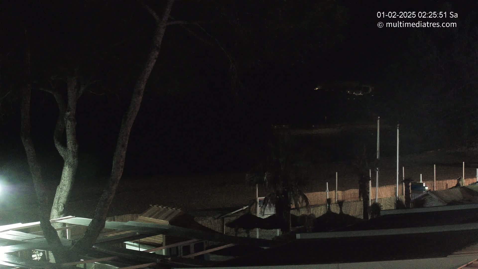A nighttime view of a dark beach with some structures and trees, under a clear, dark sky.
