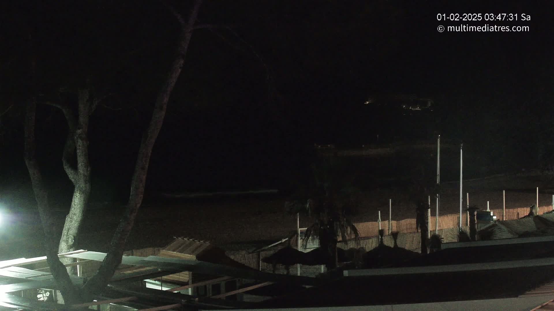 A nighttime view of a dark beach with structures and trees, under a clear, dark sky.