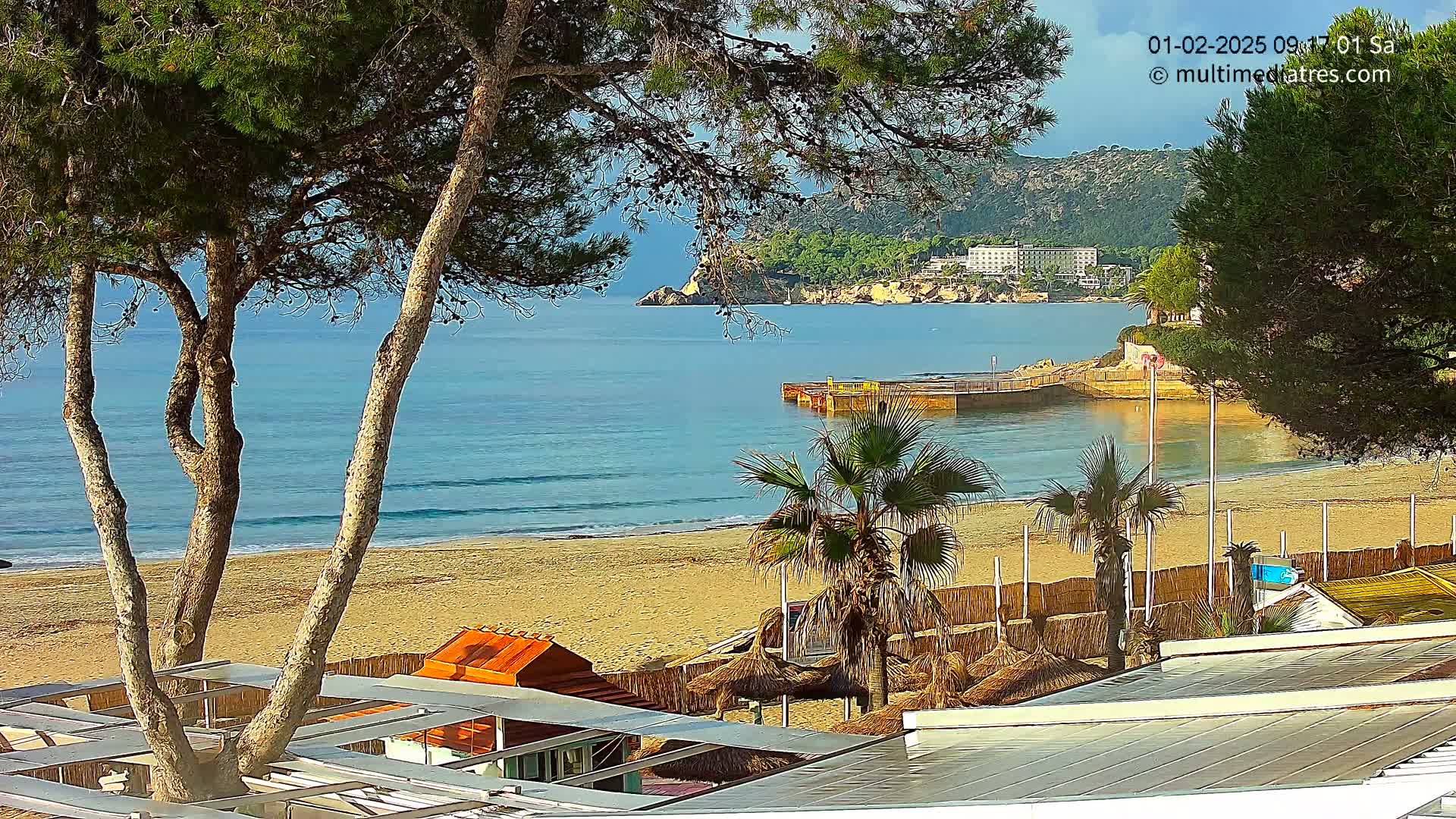 A sandy beach with calm, blue water, palm trees, and beachside structures is visible from a viewpoint above, under a mostly clear sky.