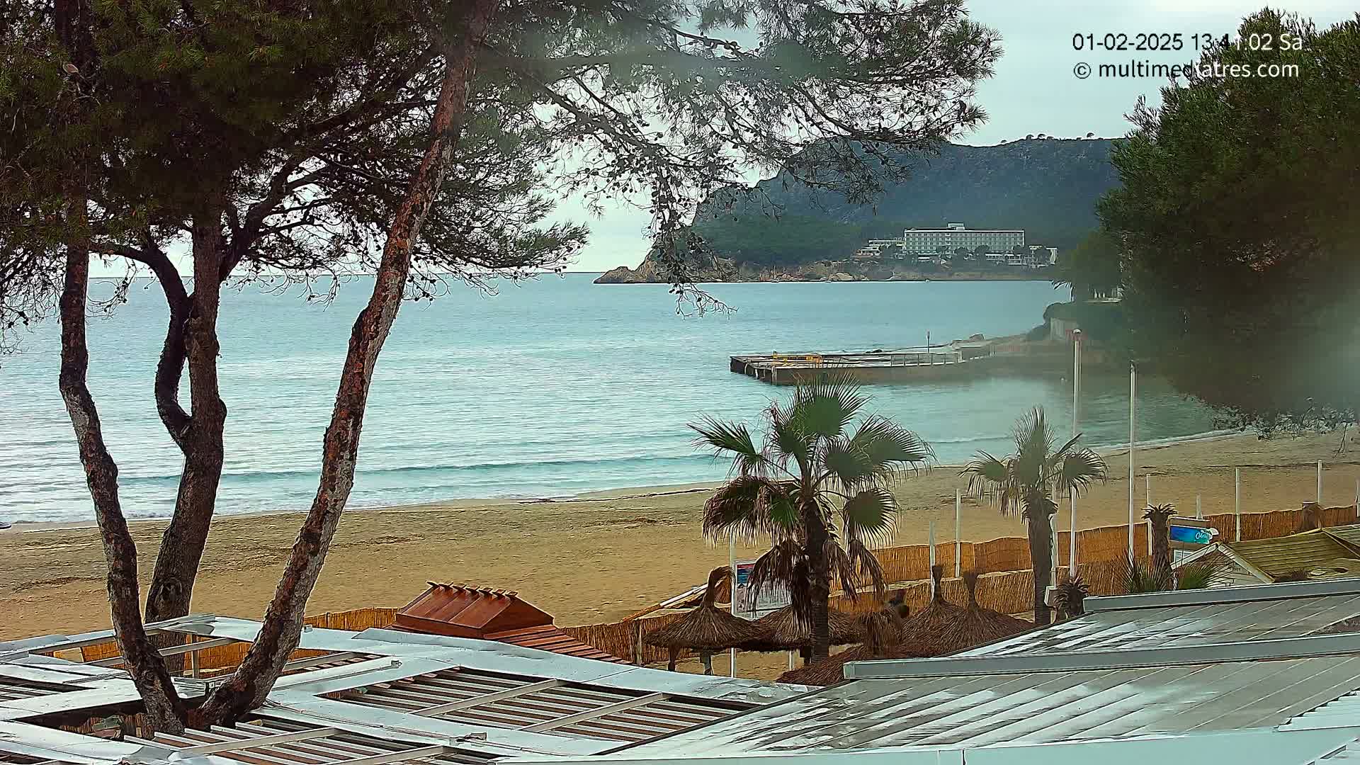 A sandy beach with calm, blue water, palm trees, and beach huts is visible under an overcast sky, with a large hotel in the distance.