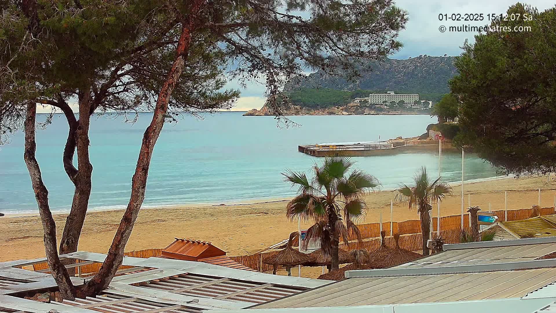 A sandy beach with calm, light-blue water, palm trees, and beachside structures is seen from an elevated vantage point on an overcast day.