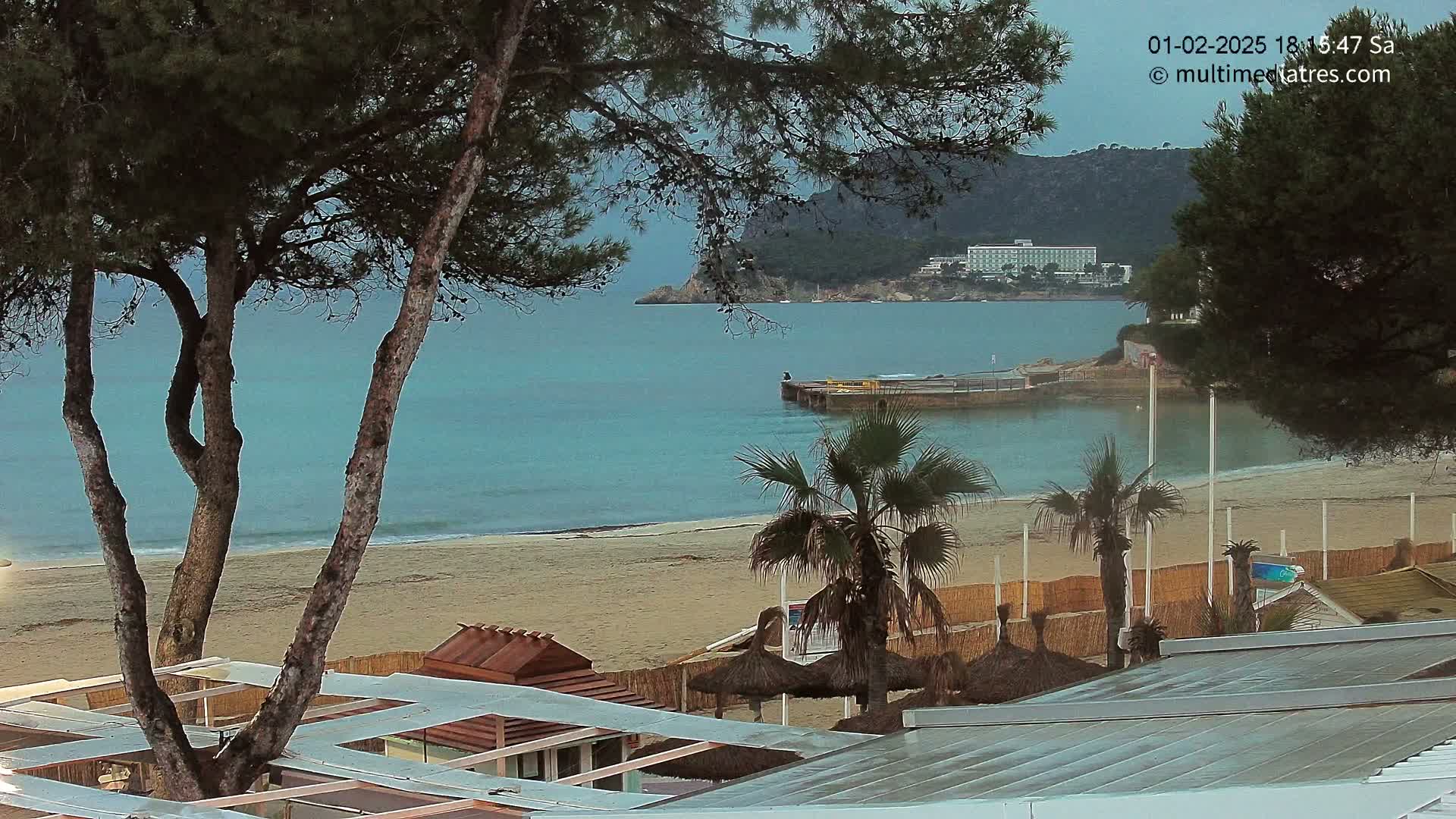 A sandy beach with calm, turquoise water is viewed from behind beachside structures and trees, under an overcast sky.