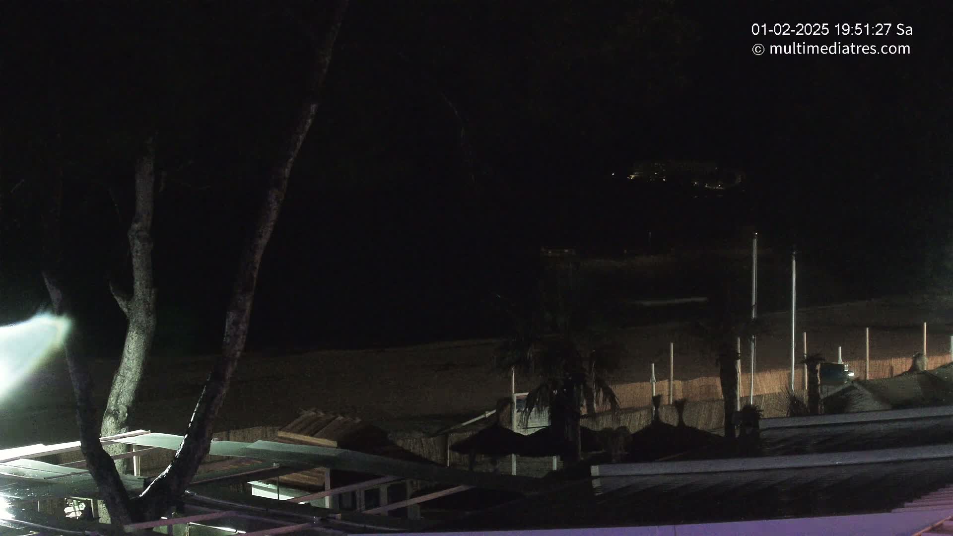 A nighttime view of a dark beach with some structures and palm trees, under a clear sky.