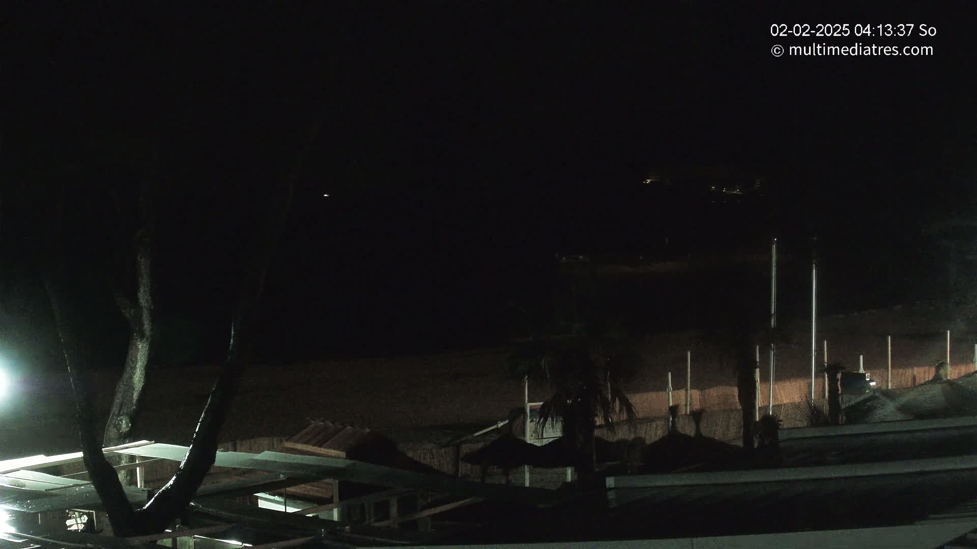 A nighttime view of a dark beach with structures and trees, illuminated by a single light source.