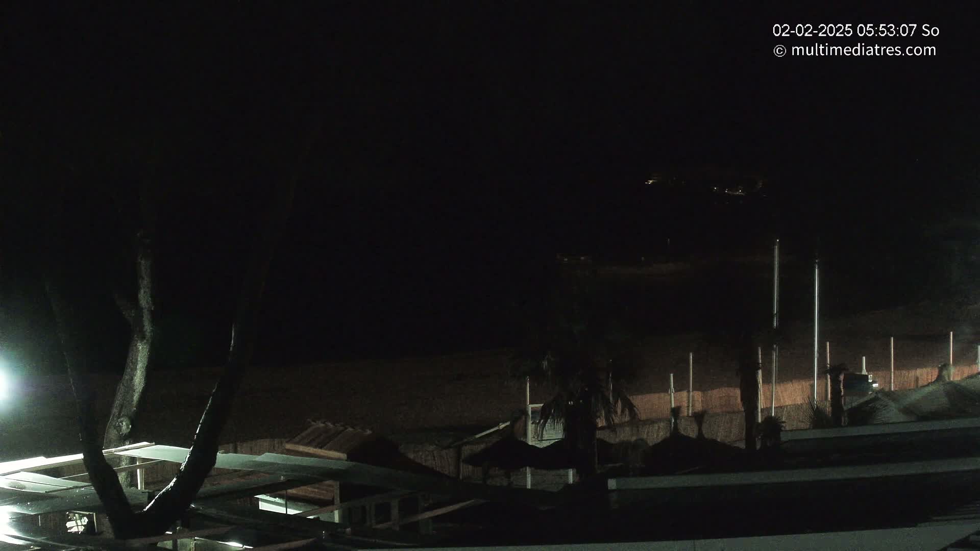 A nighttime view of a dark beach with dimly lit structures and a few palm trees, under a clear, dark sky.