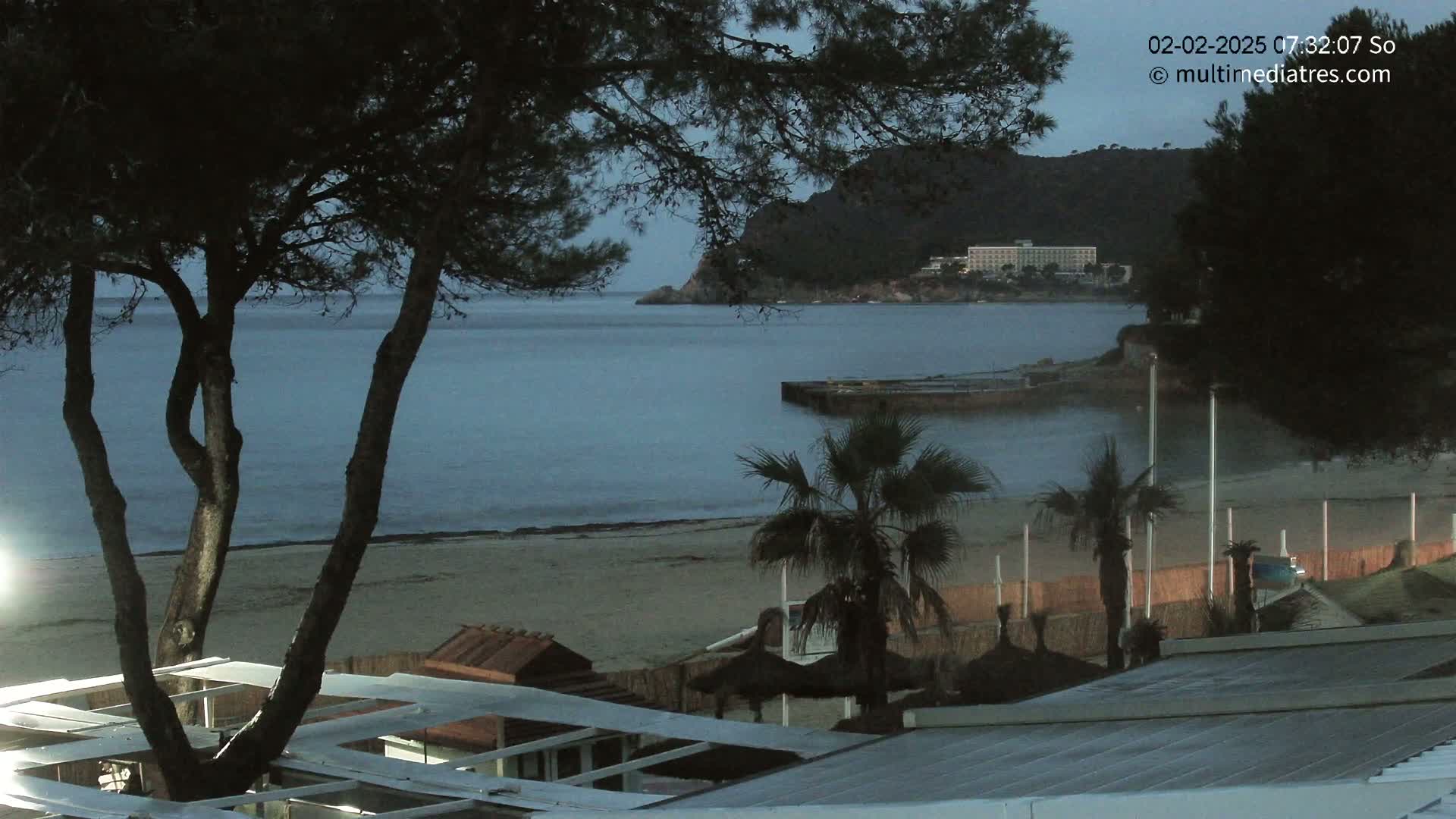 A calm, twilight ocean scene shows a sandy beach with palm trees, beach structures, and a distant hotel, all under a dark sky.