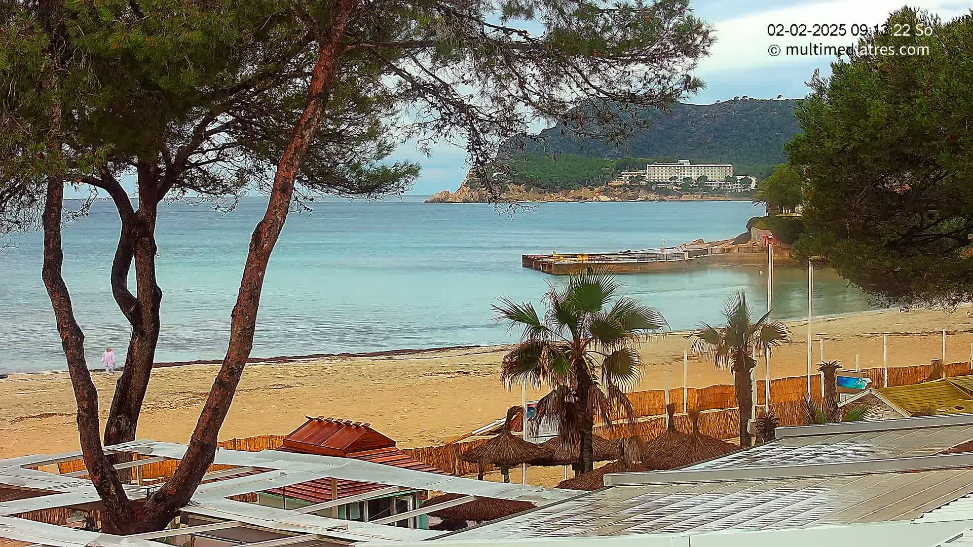 A sandy beach with calm, light-blue water, palm trees, and beach structures is seen from above, with a hotel and hills visible in the distance under a mostly cloudy sky.