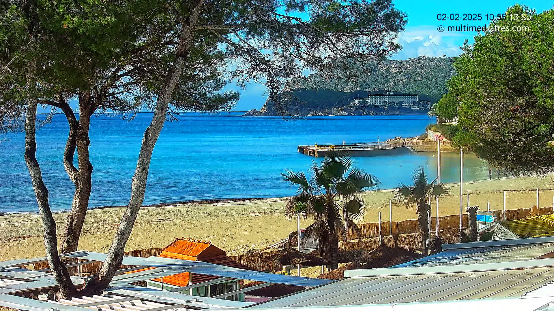 A sandy beach with turquoise water, palm trees, and beachside structures is visible under a sunny sky, with a distant hillside and buildings in the background.