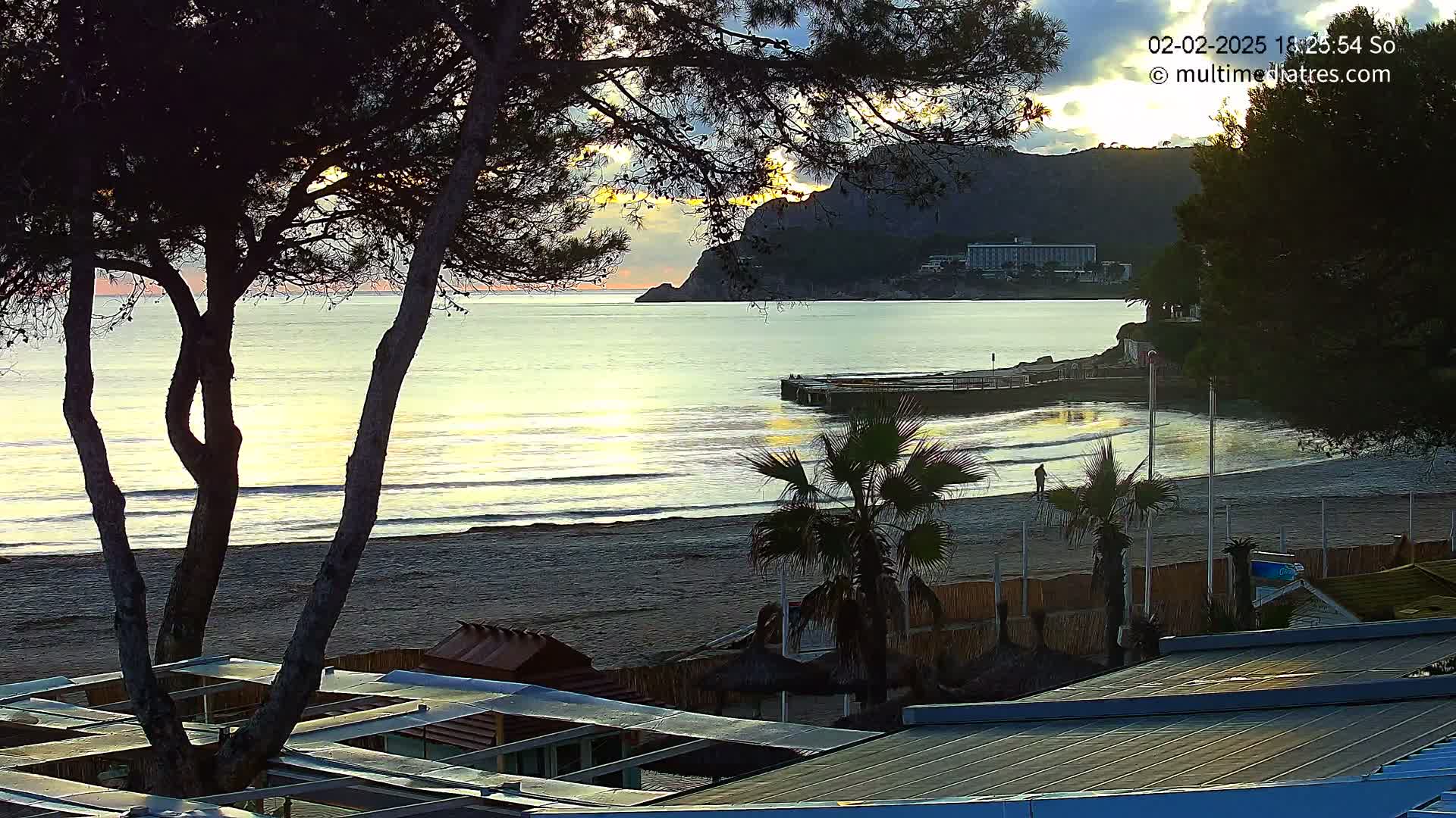 A sandy beach with calm waves and a few palm trees at sunset, overlooked by a hillside with buildings and silhouetted trees in the foreground.