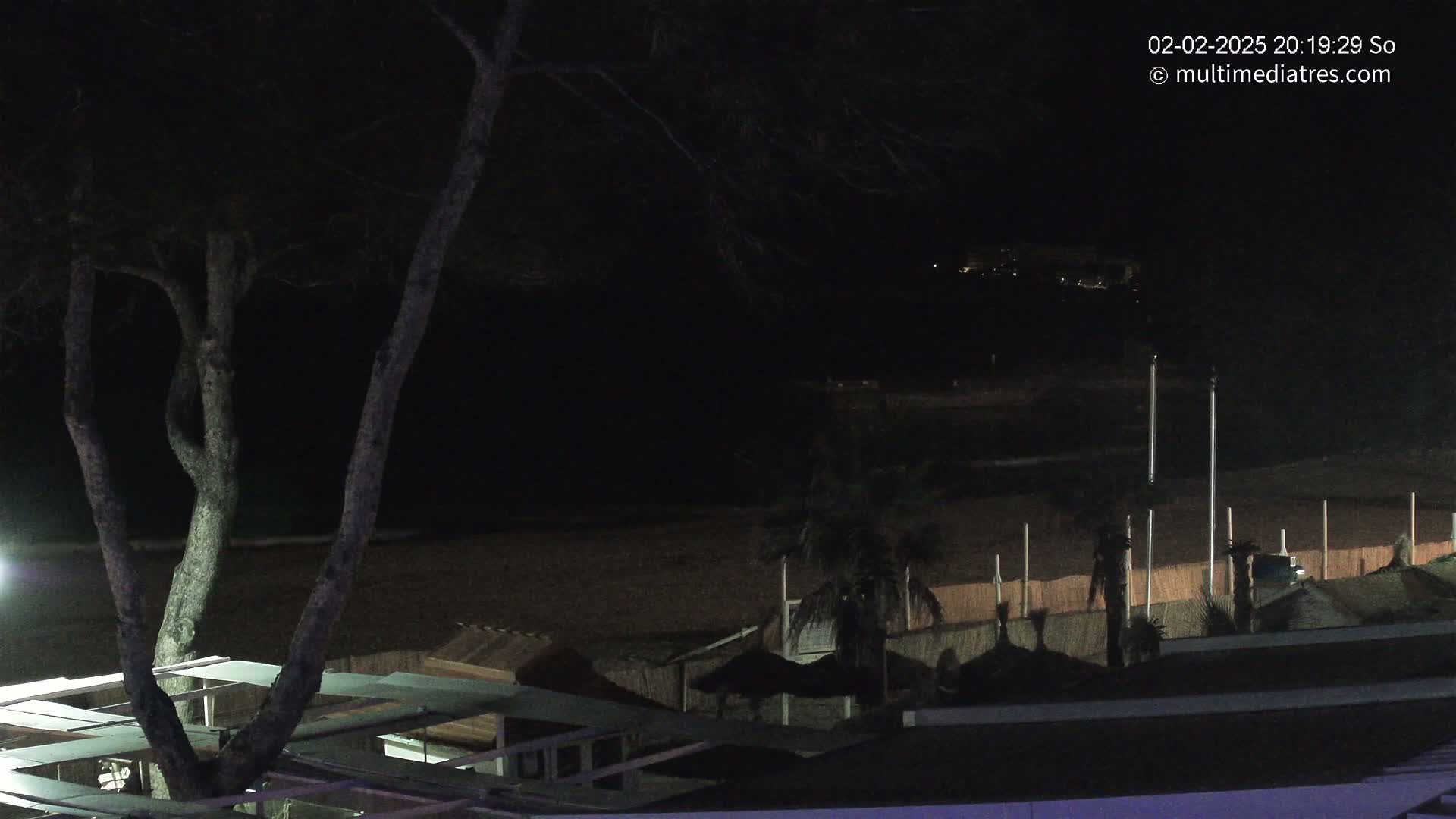 A dark, nighttime beach scene shows a sandy shore with several palm trees and beach structures under a mostly clear sky.