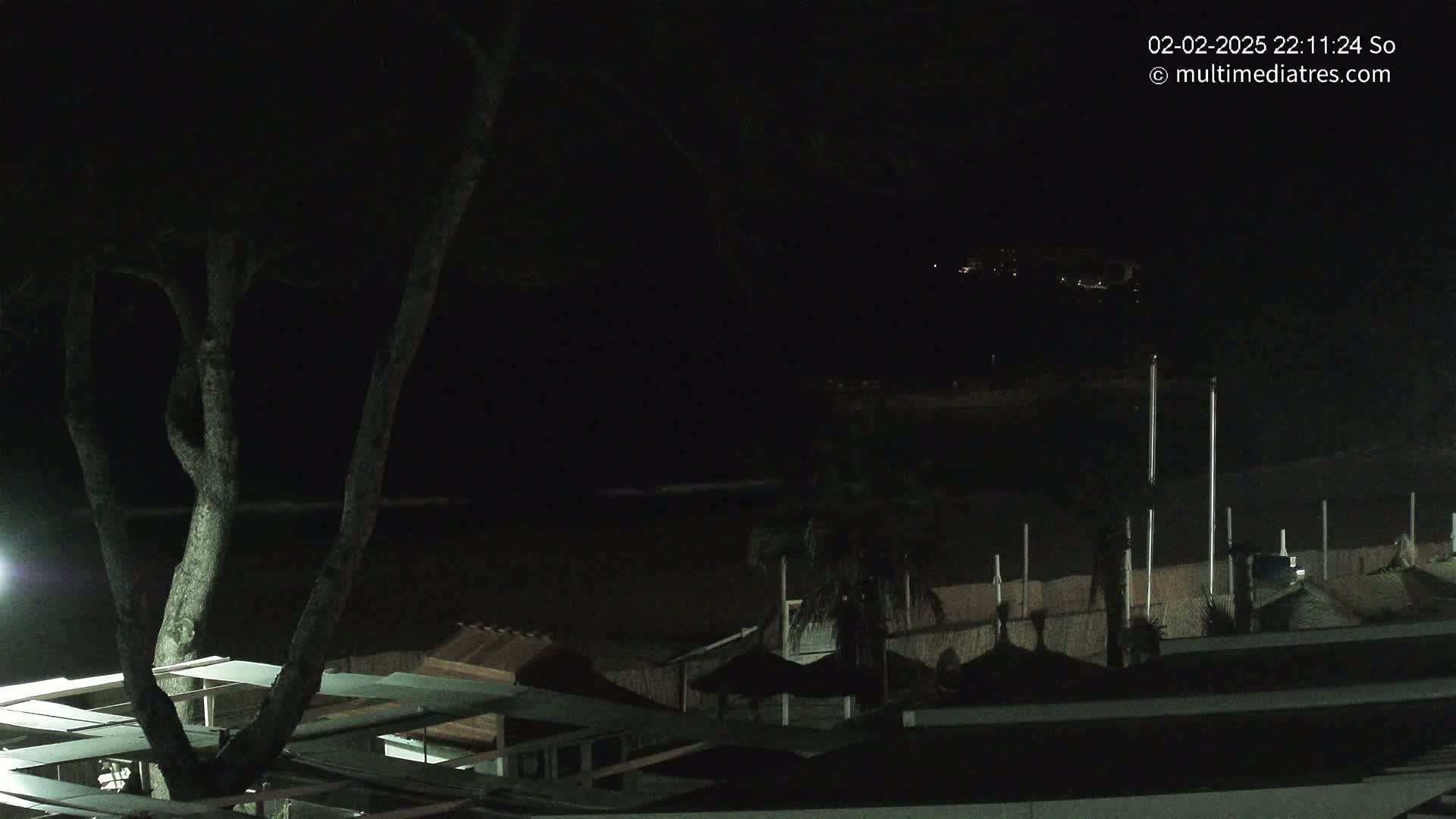 A nighttime view of a dark beach with a few dimly lit structures and a tree, under a clear, dark sky.