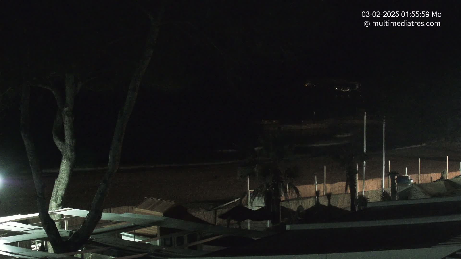 A dark nighttime beach scene shows a mostly empty sandy shore with a few palm trees and beach huts under a clear, dark sky.