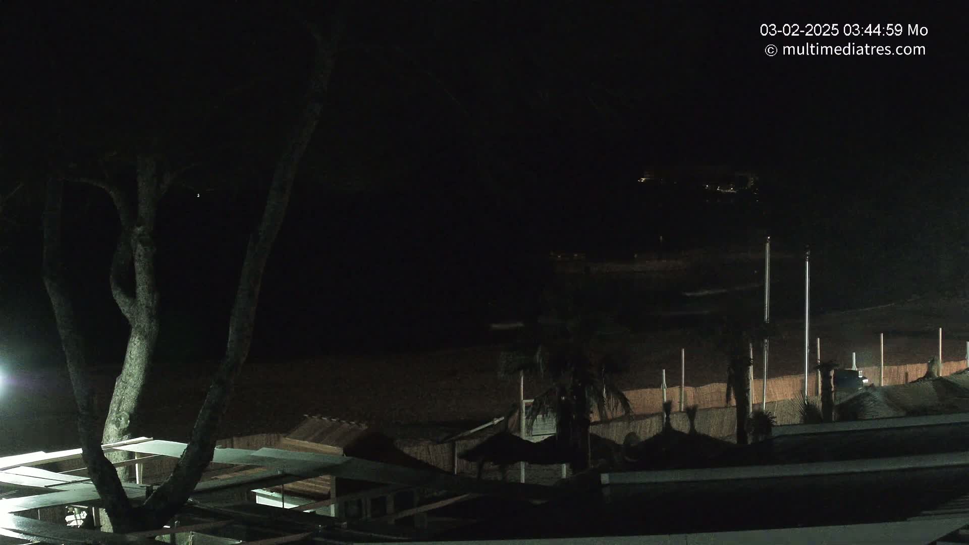 A dark nighttime view of a beach with some structures, trees, and a dimly lit shoreline.