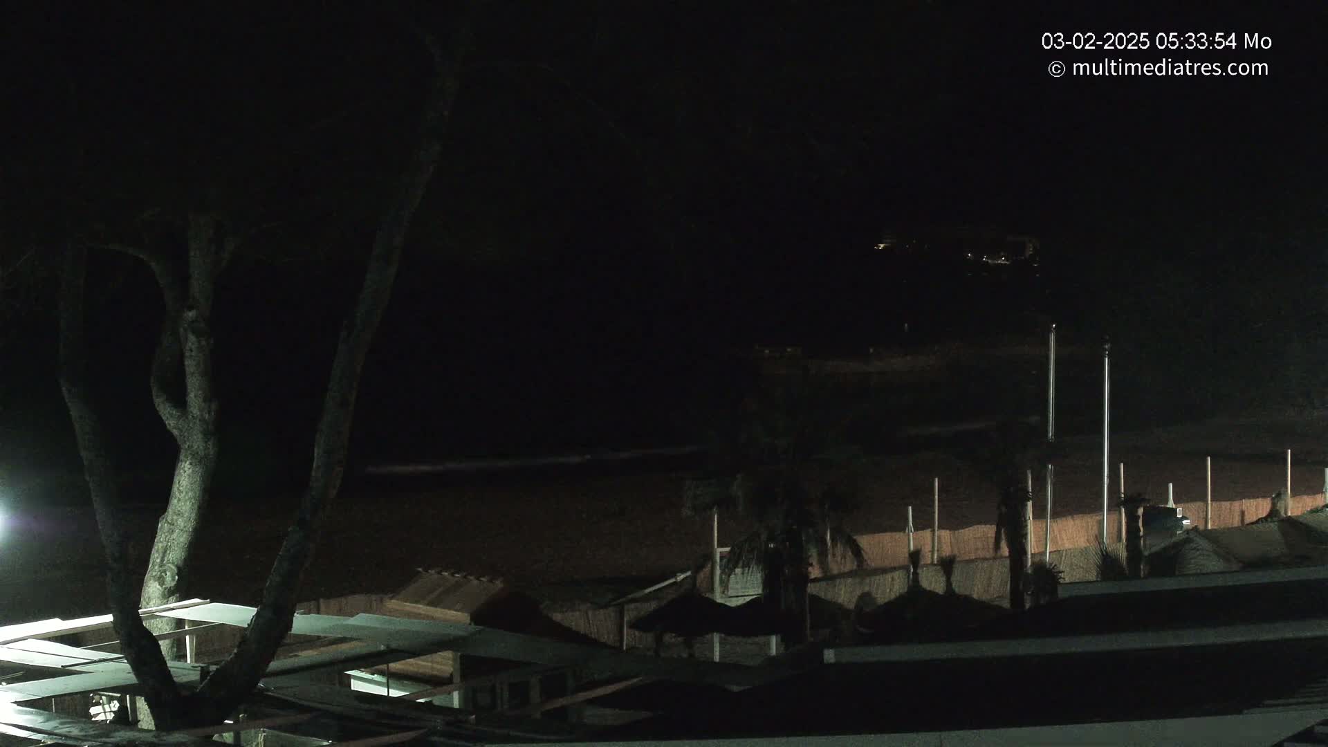 A dark nighttime beach scene shows a mostly obscured beachfront with some structures and palm trees, under a clear, dark sky.