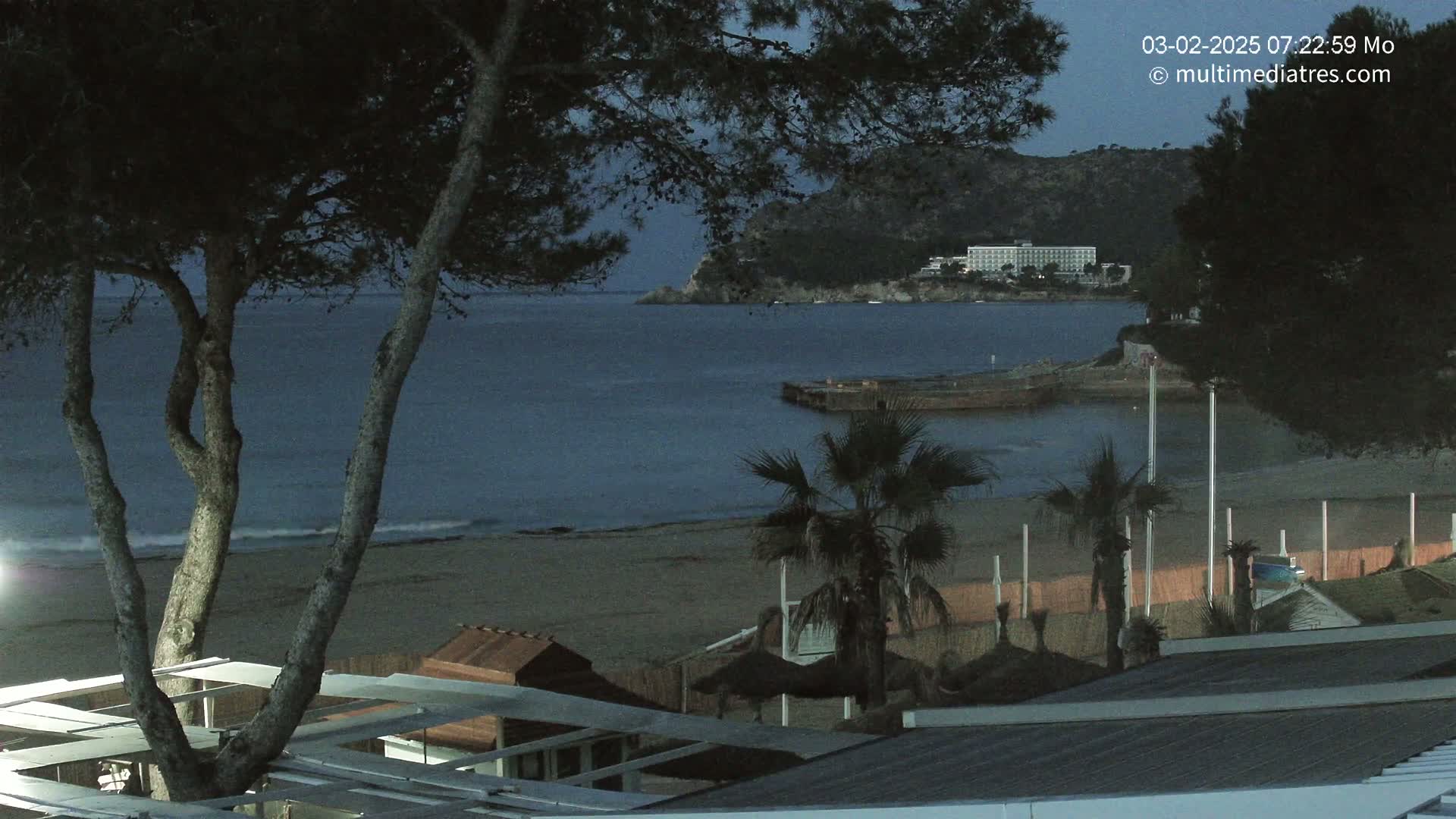 A sandy beach at dusk, with palm trees, a hotel in the distance, and calm water under a dark sky.