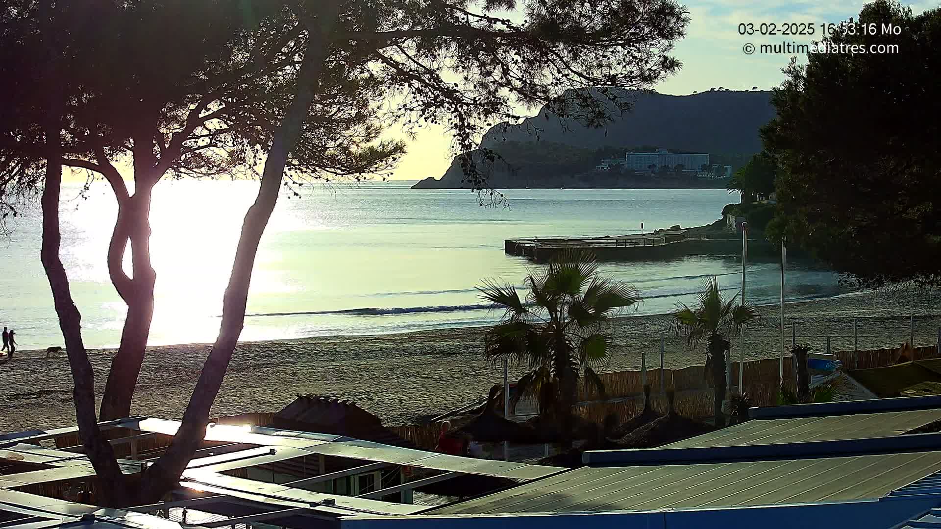 A sandy beach with calm waves at sunset, seen from behind trees and beachside structures, with a distant mountain range visible.