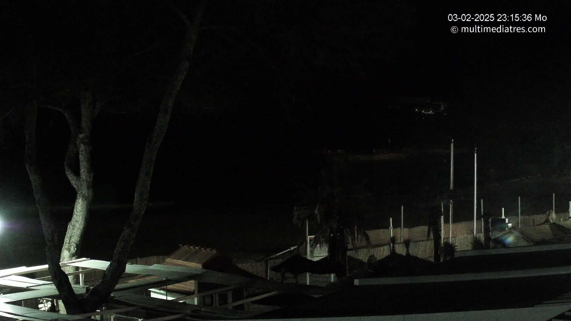 A dark nighttime scene overlooking a beach with several structures, a tree, and dimly lit water.