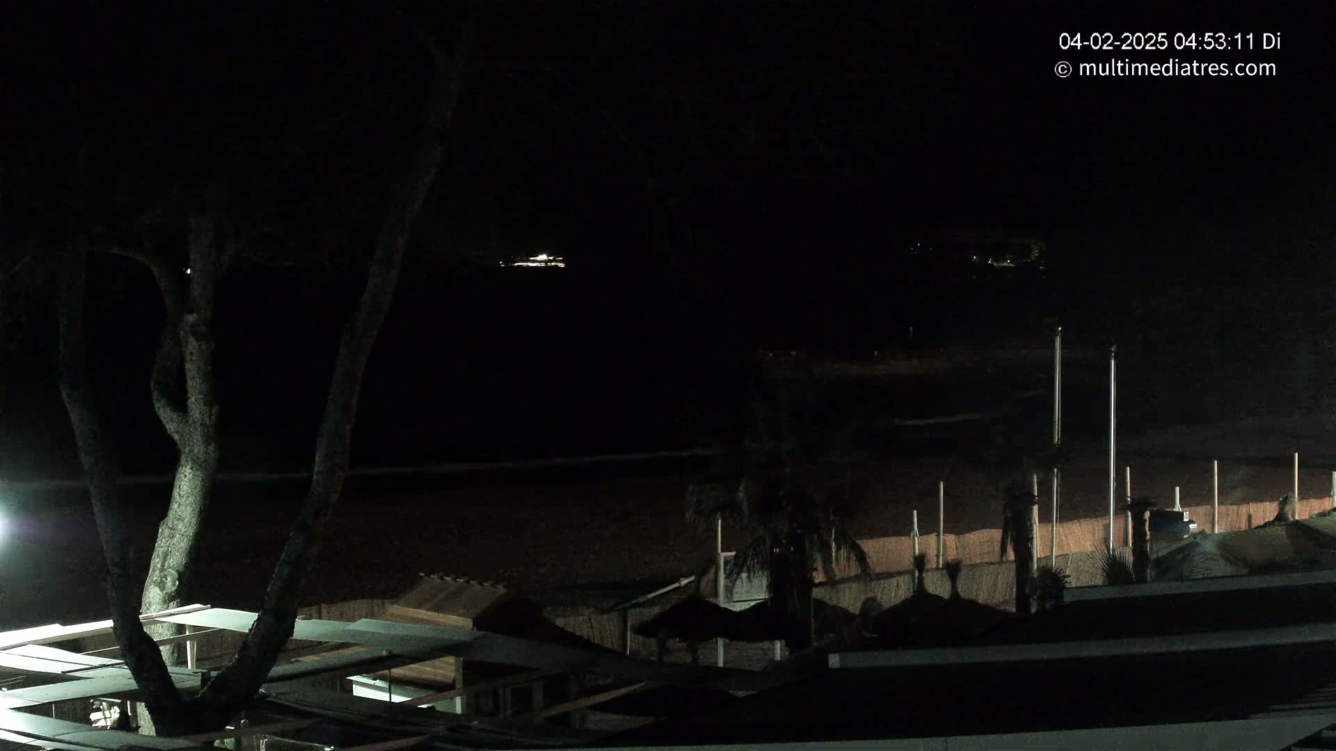 A dark nighttime scene overlooking a beach with dimly lit structures and a vessel visible in the distance on the water.