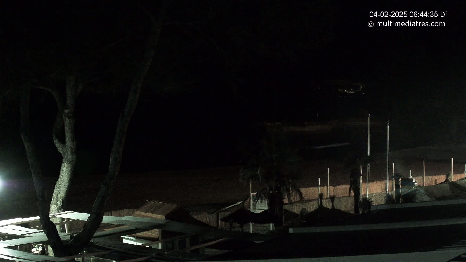A nighttime view of a beach at night, showing dark sand, a few palm trees, and beach structures partially illuminated under a clear, dark sky.