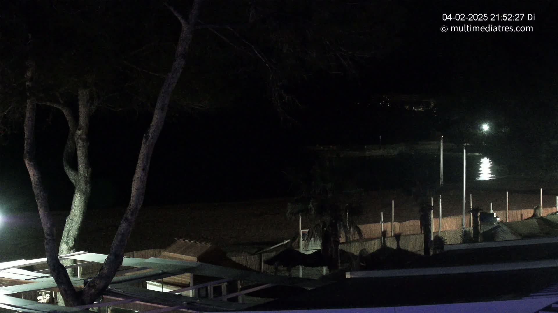 A nighttime view of a beach at night, showing dark trees, a calm body of water reflecting light, and beach structures under a dark sky.
